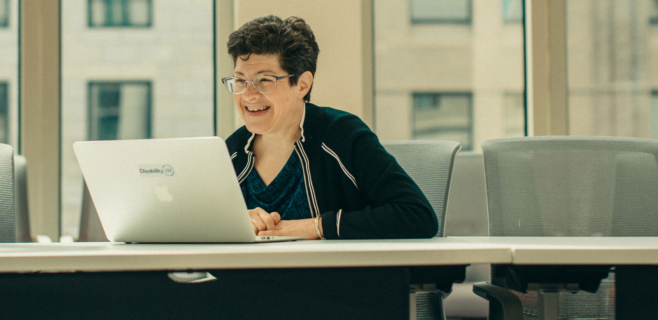 A woman sits at a conference table, using a laptop, smiling at the camera.