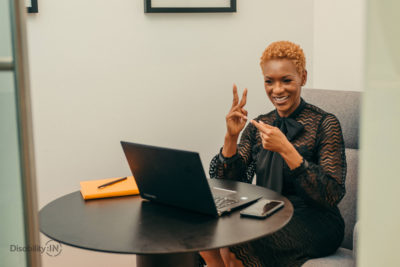 Woman of color smiles and signs ASL to a computer as if it's a video conference.