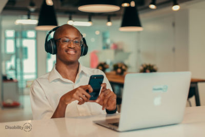 Man sits in front of laptop with headphones on smiling while also looking at his phone.