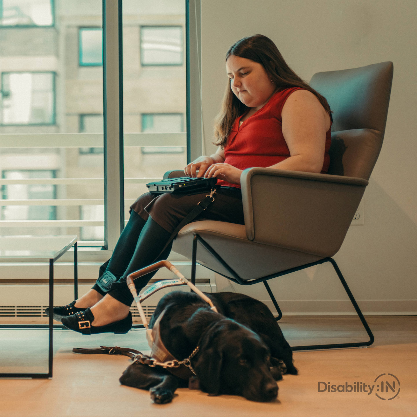 Woman sits on chair taking notes on braille notetaker. Guide dog sits on floor beside her.