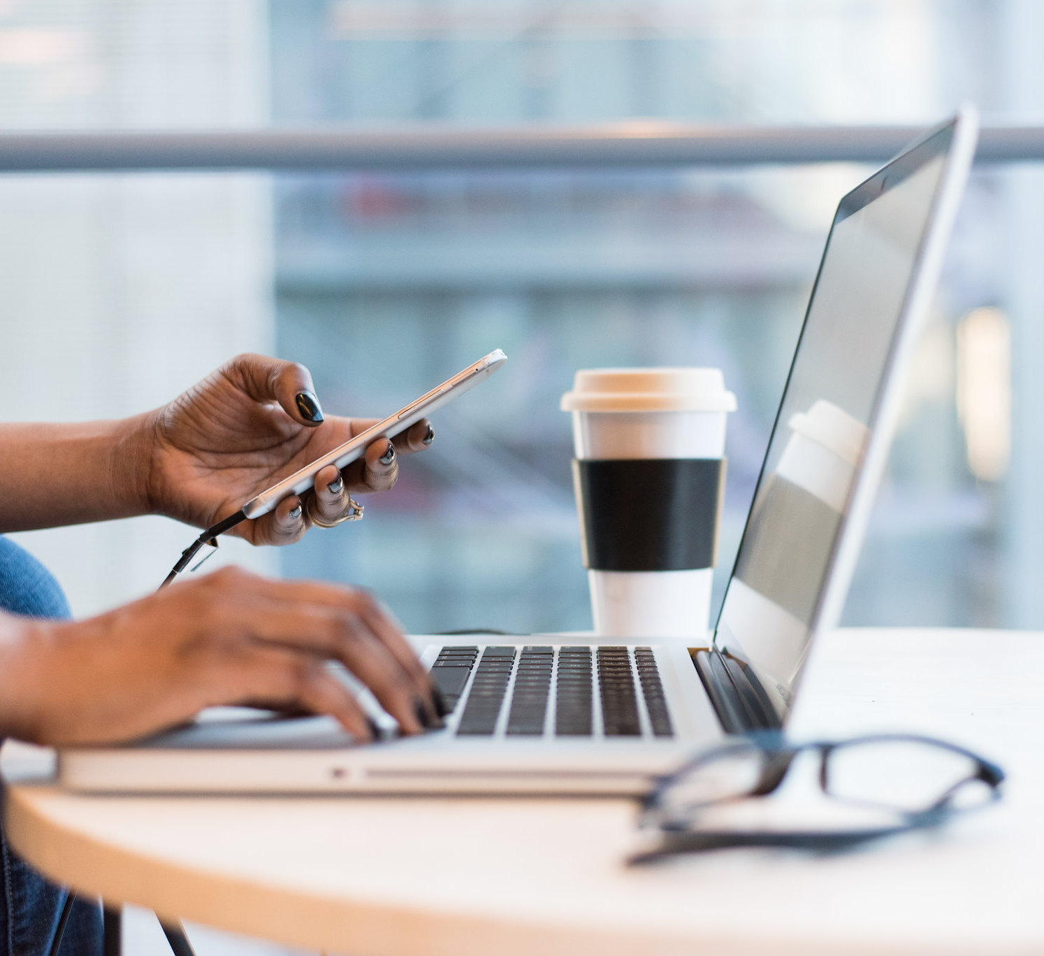 Up close of Black woman working on her laptop while holding her phone near a cup of coffee