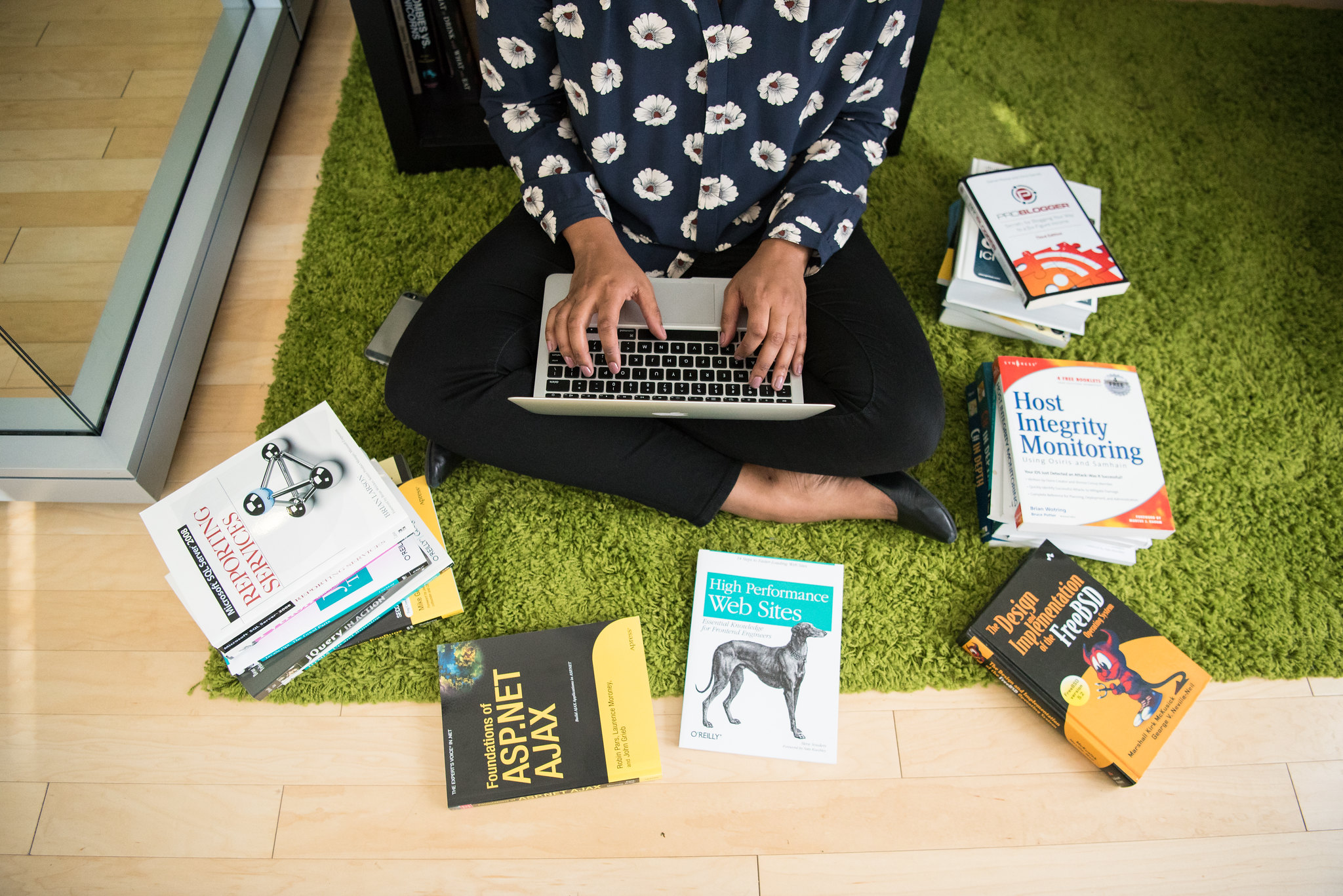Woman of color sits on floor working on laptop surrounded by books