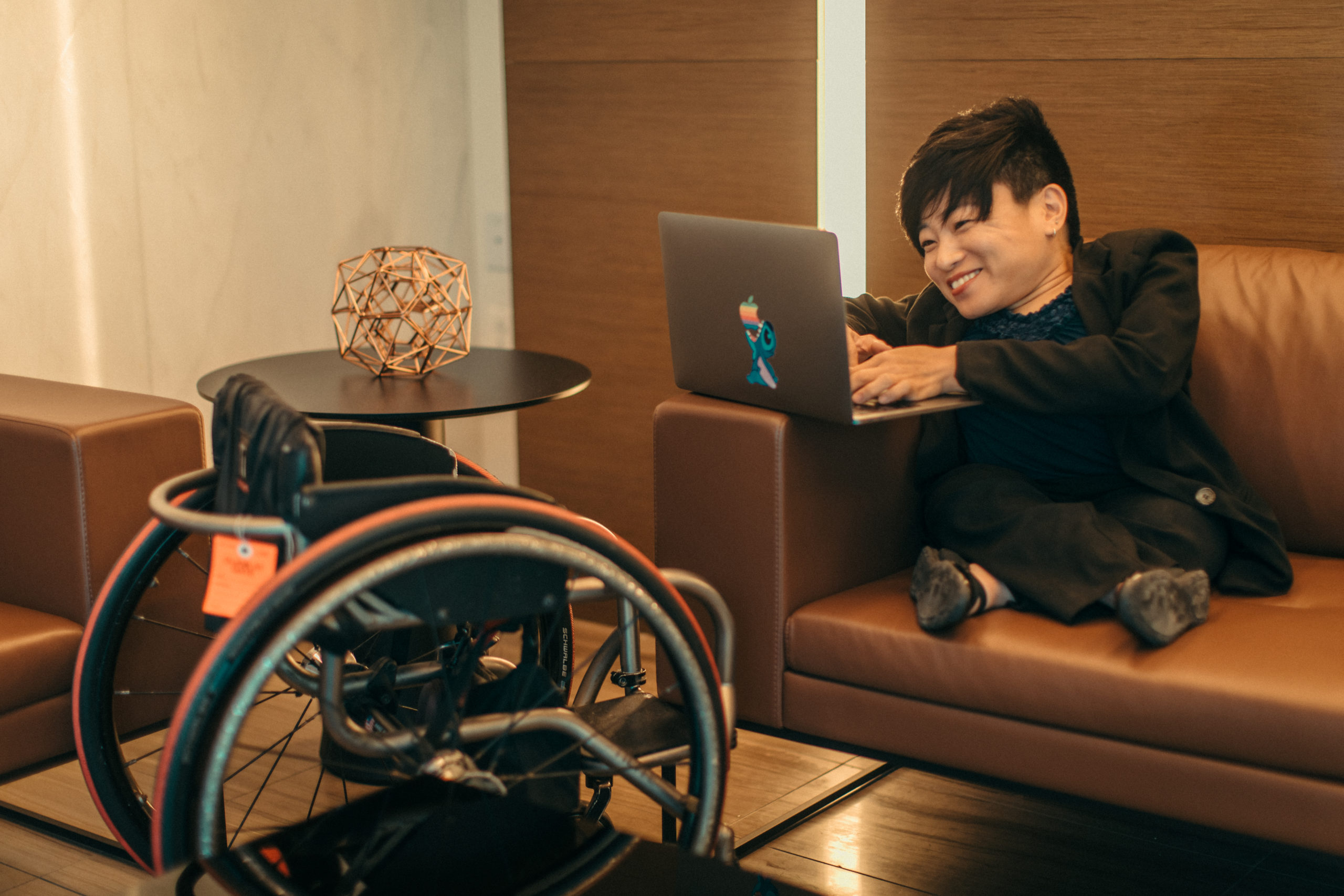Woman sits on couch working on laptop with wheelchair across from her. She is smiling and is in a corporate setting/lounge.