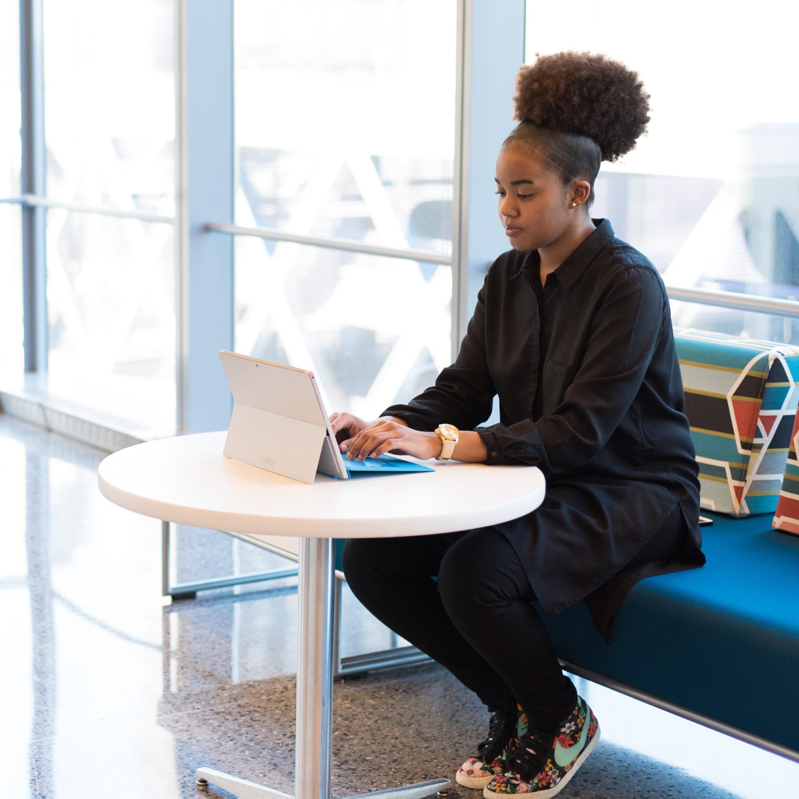Black woman with black jacket and colorful sneakers sits at a bench working on her laptop.