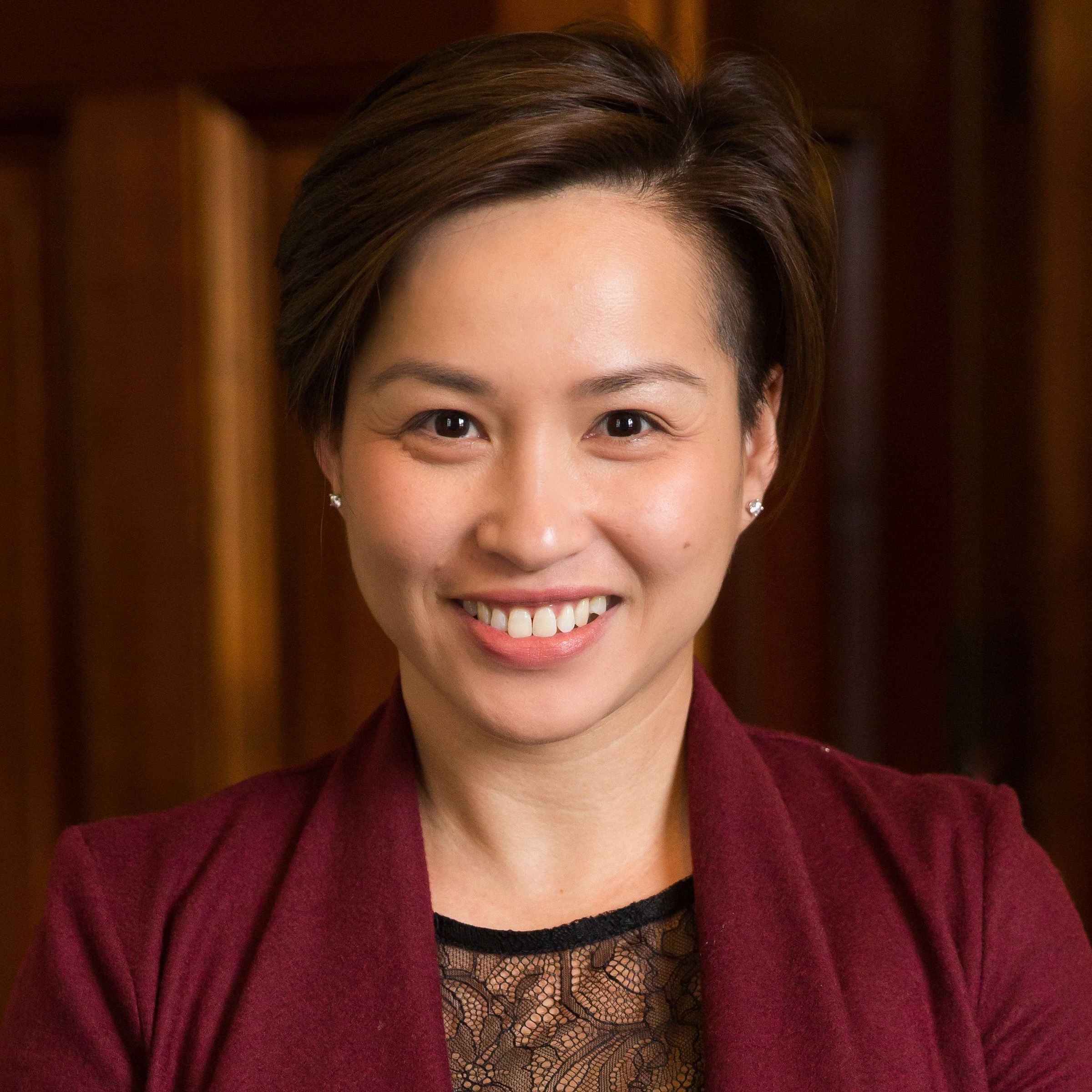 Asian woman with short hair and red blazer smiles against dark background.