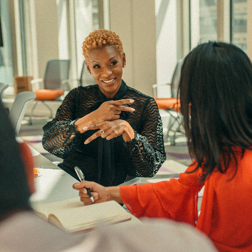 A black woman with short hair and black blouse signs to her colleague in red top while sitting at a conference table.