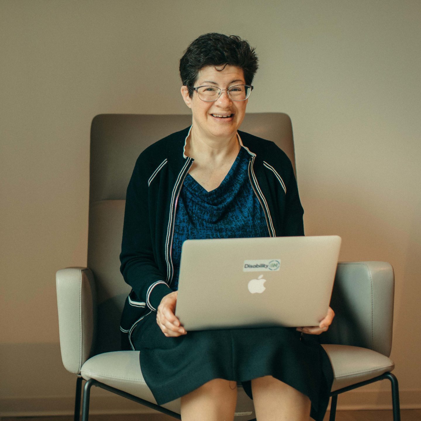 White woman with disability with short dark hair and dress with jacket while smiling and holding laptop in her lap.