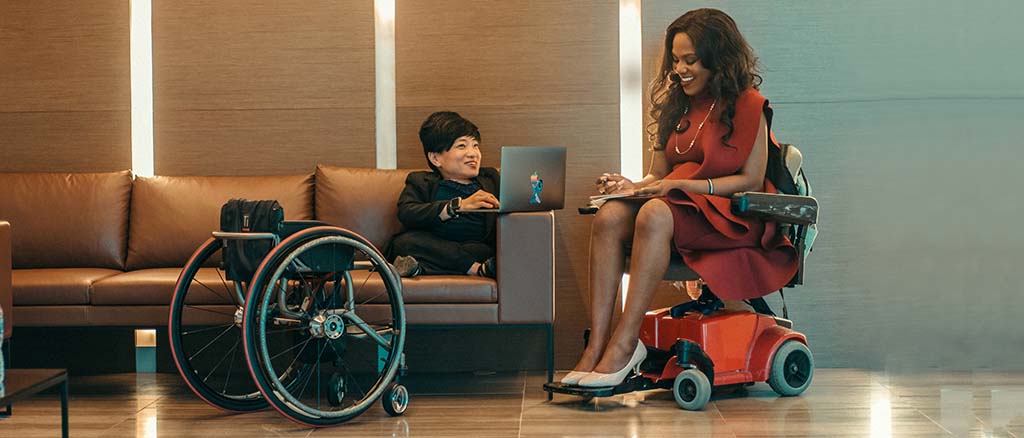 Two diverse women with wheelchairs gather together near sofa and smile and take notes on notepad and laptop.