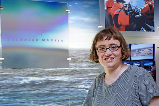 A white woman with glasses and short hair smiles next to Lockheed Martin signage.