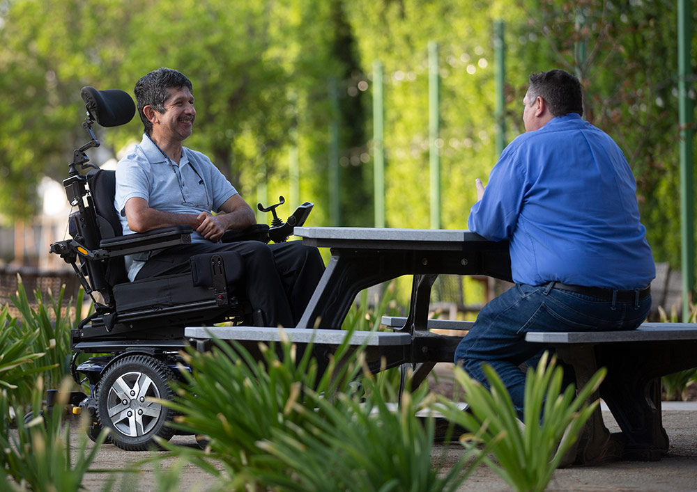 Two people with disabilities chat at an outdoor bench and table.