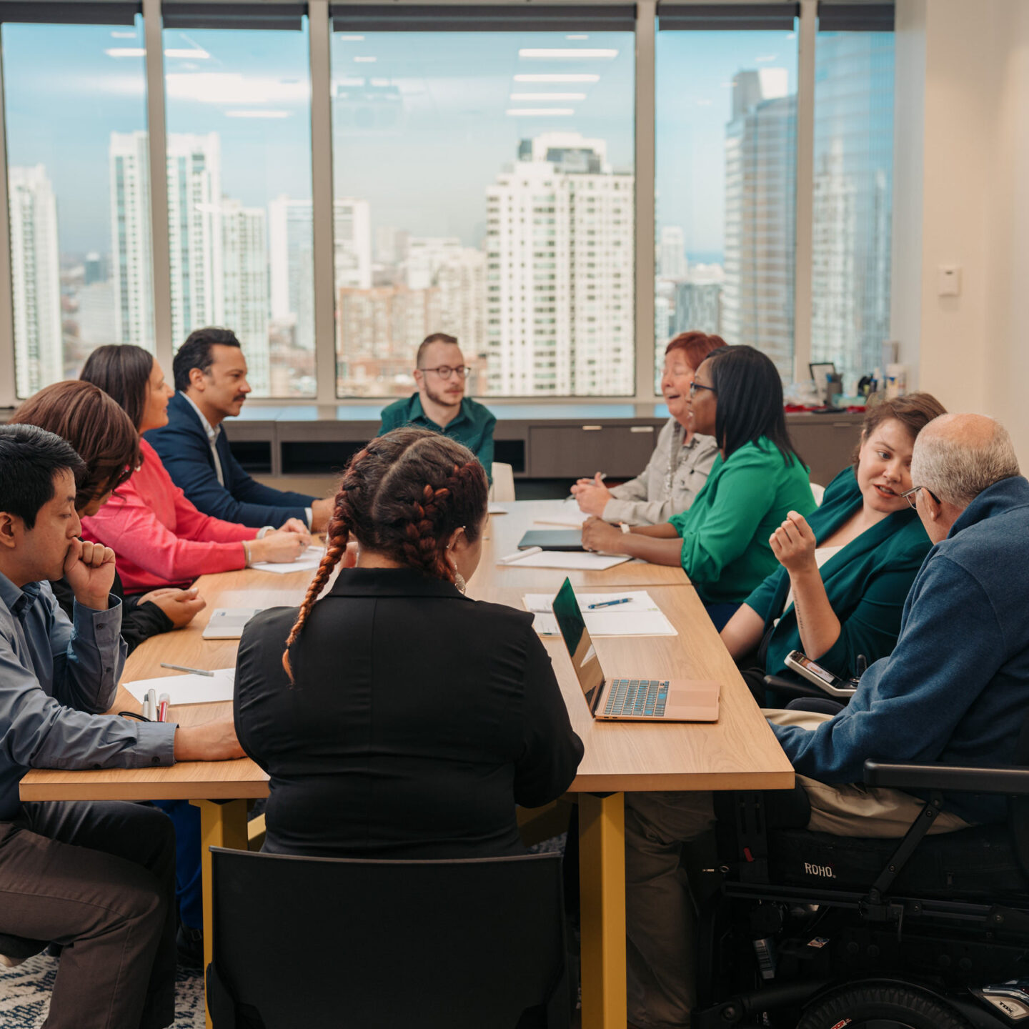 A group of diverse professionals representing various disability identities gather in a meeting around a conference table. Windows overlook a city skyline.