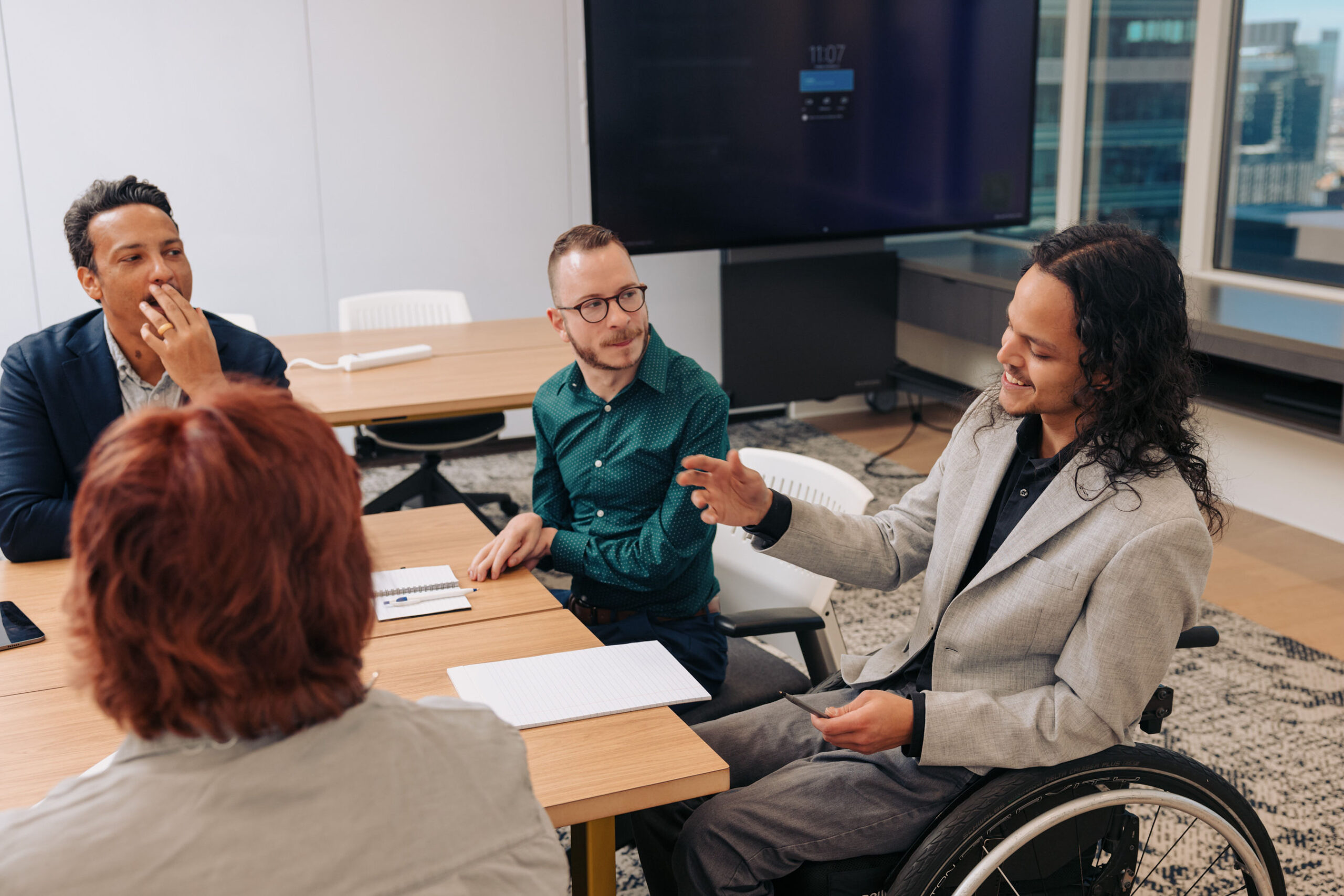 A young man who uses a wheelchair speaks to his colleagues during a meeting. They are seated around a conference table scattered with papers.