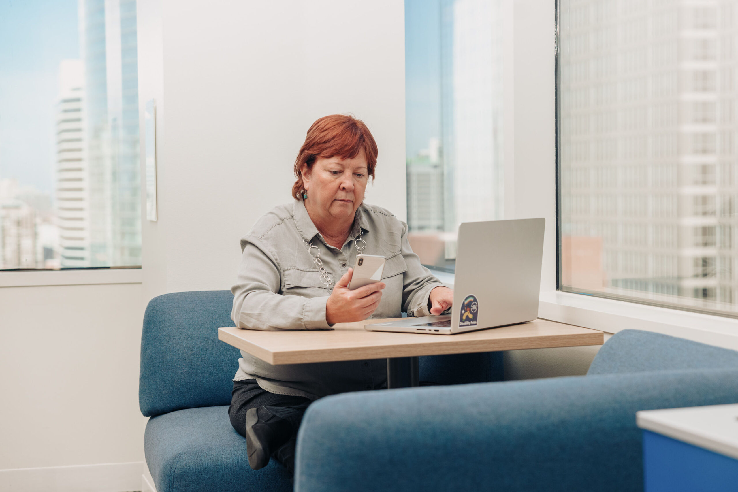 An older woman works in an office on her laptop. She holds a phone in her other hand.