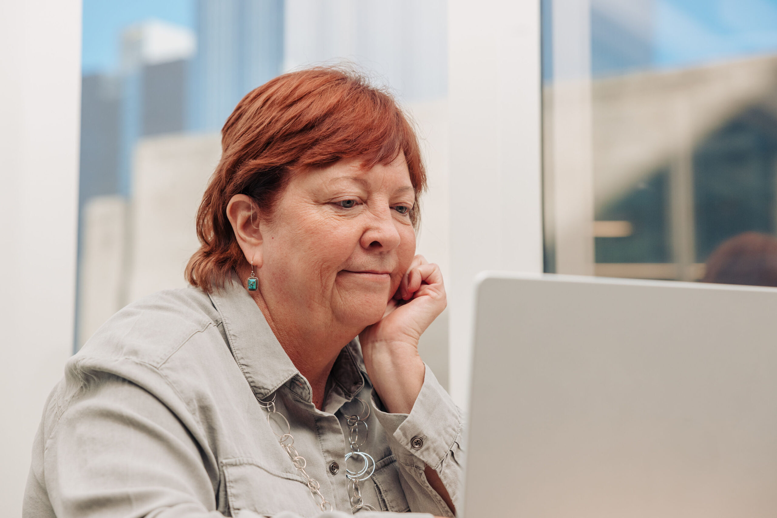 An older woman works on her laptop in an office. Her red hair is tucked behind her ear to reveal her hearing aid.