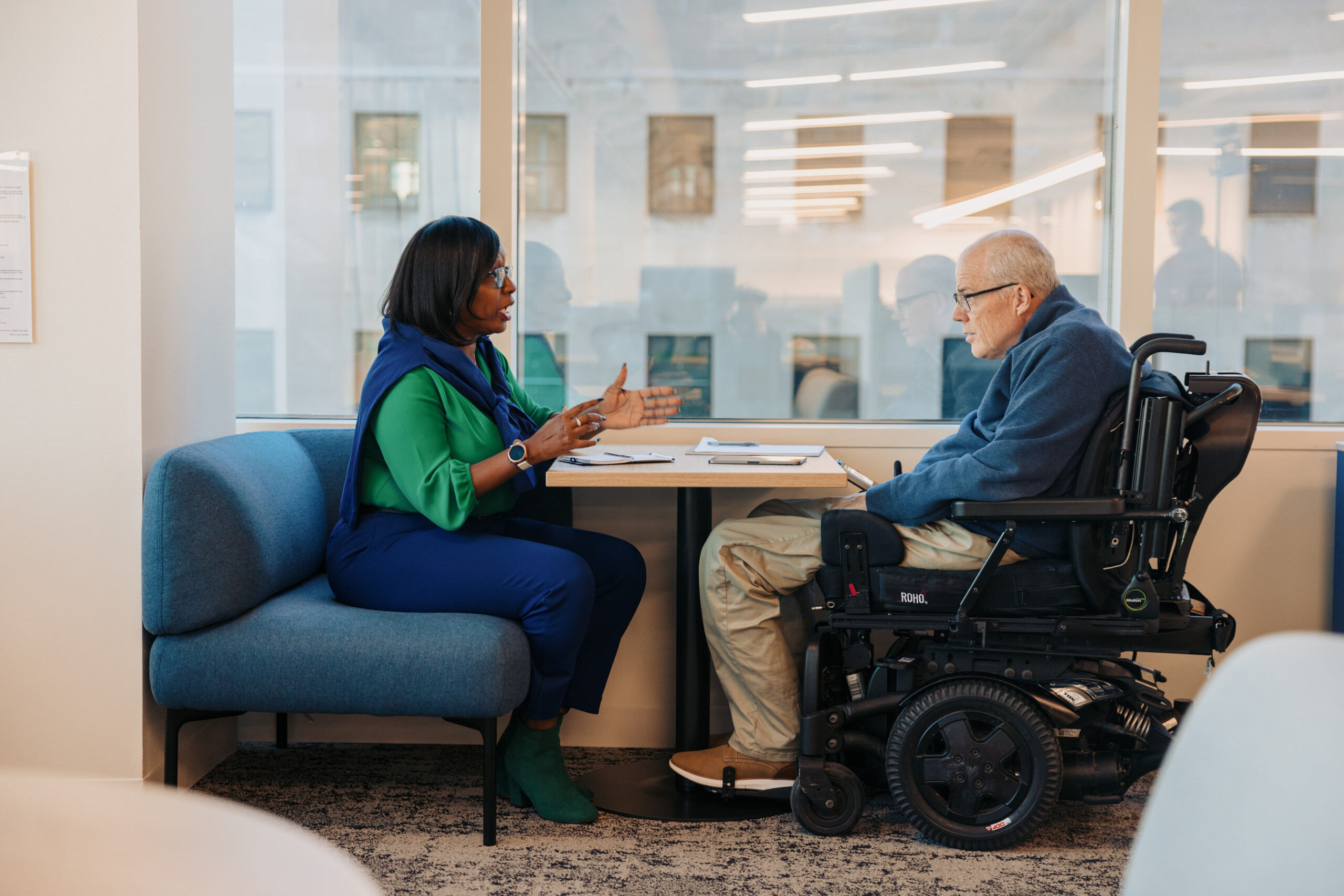 Two colleagues sit across from one another in a meeting. One uses a power wheelchair.
