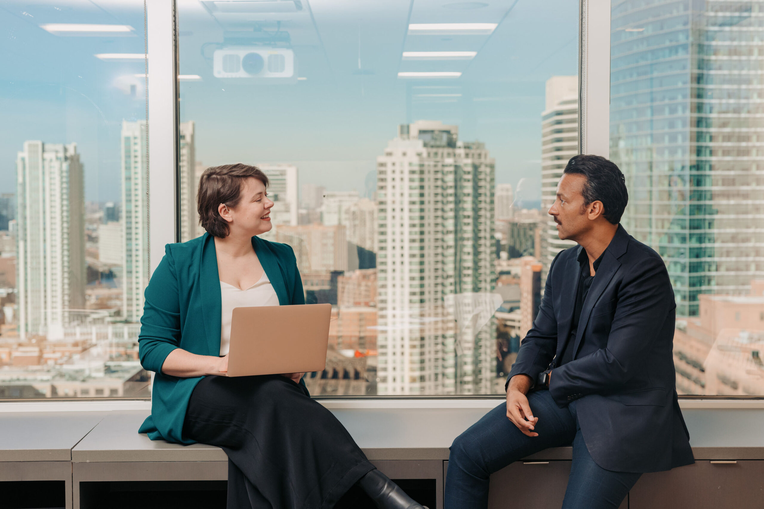 Two professionals sit on a window-seat in an office that overlooks a city skyline. One wears a hearing aid and holds a laptop in their lap.