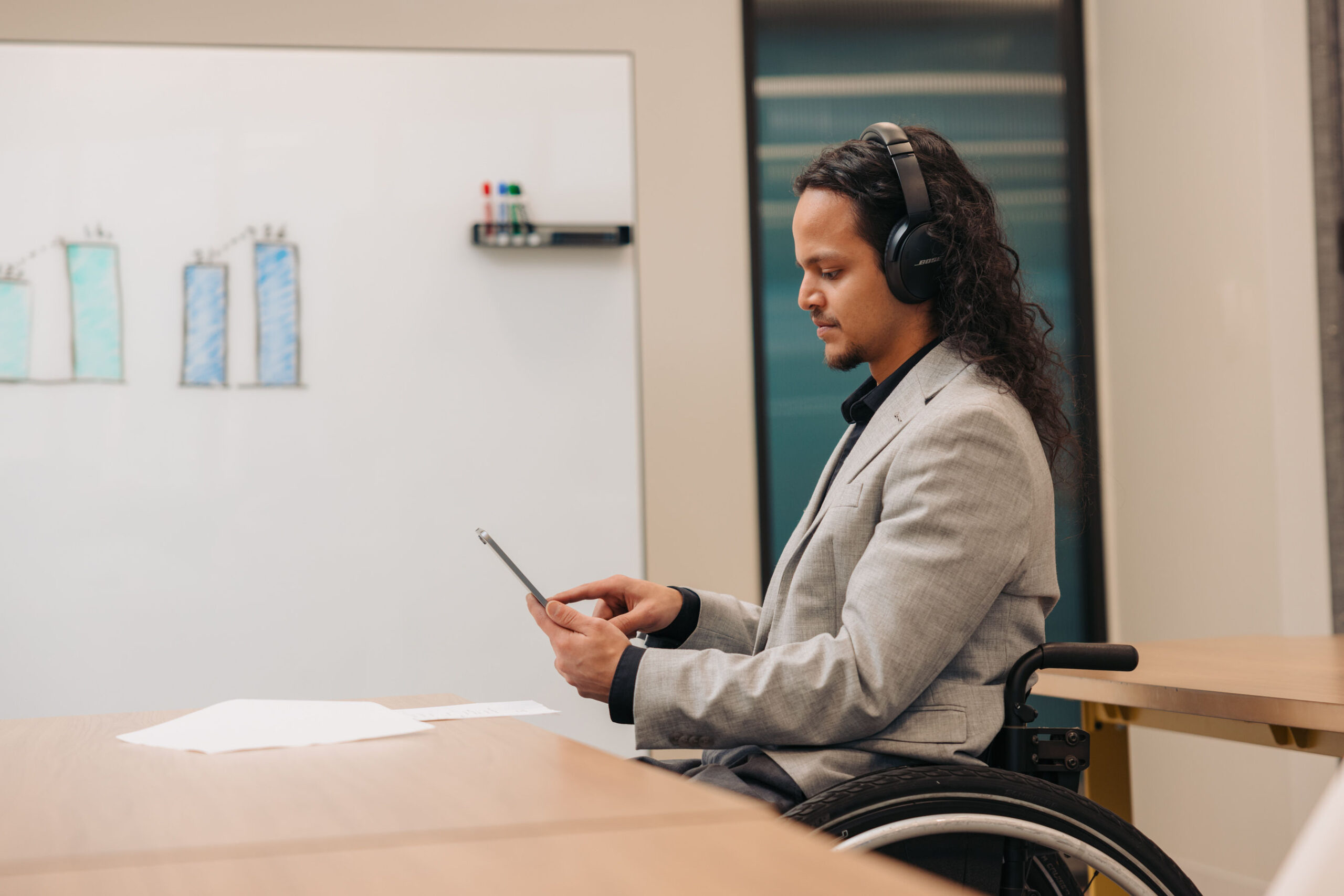 A young man using a wheelchair works in a conference room. He wears noise-cancelling headphones and types on a tablet.