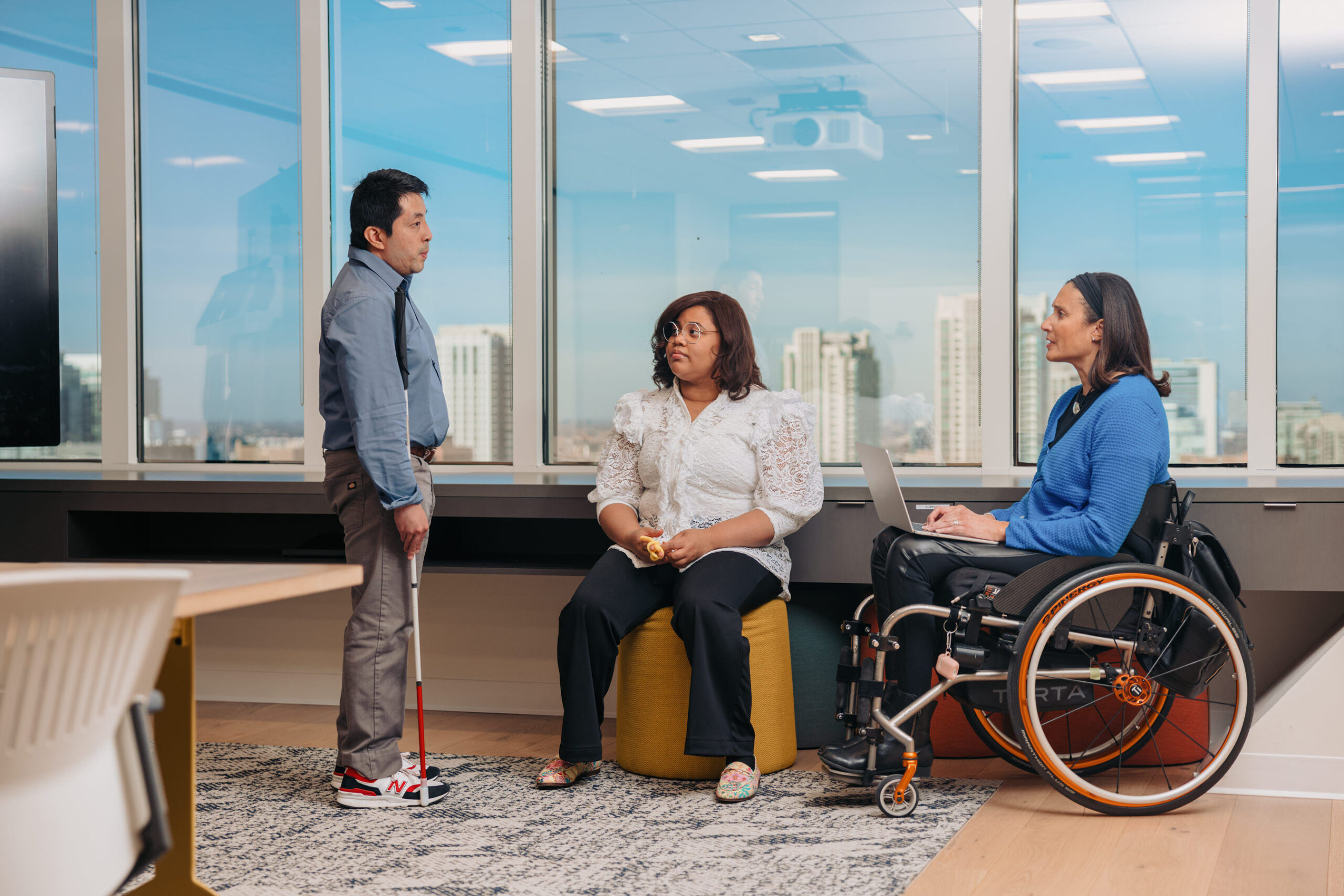 Three colleagues converse in a meeting room. One stands with his white cane in one arm. Beside him, a young woman sits holding a fidget toy next to a woman who is using a wheelchair.