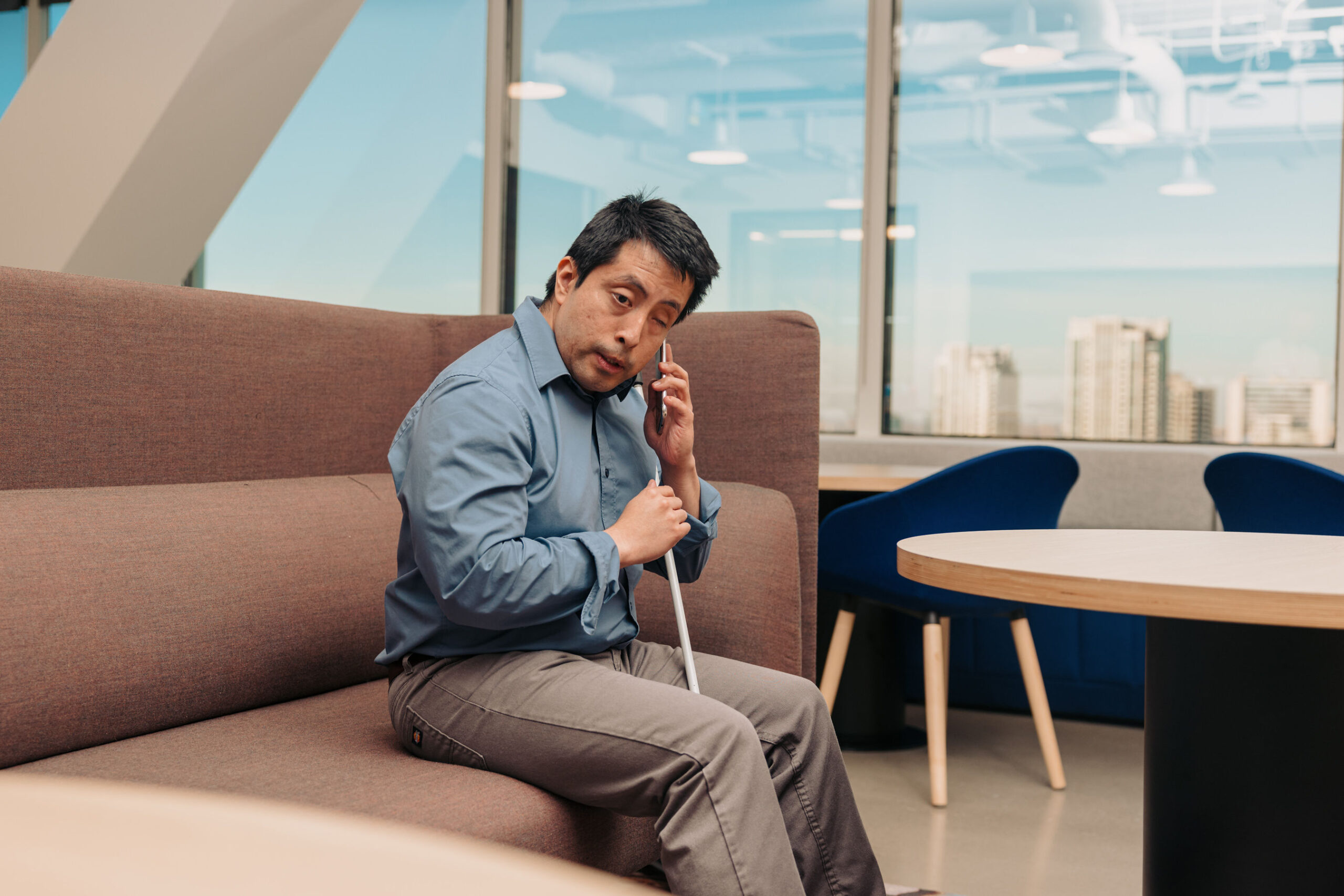 A man who is blind sits in an office as he listens to the text to speech on his phone. He sits with his white cane resting between his legs.