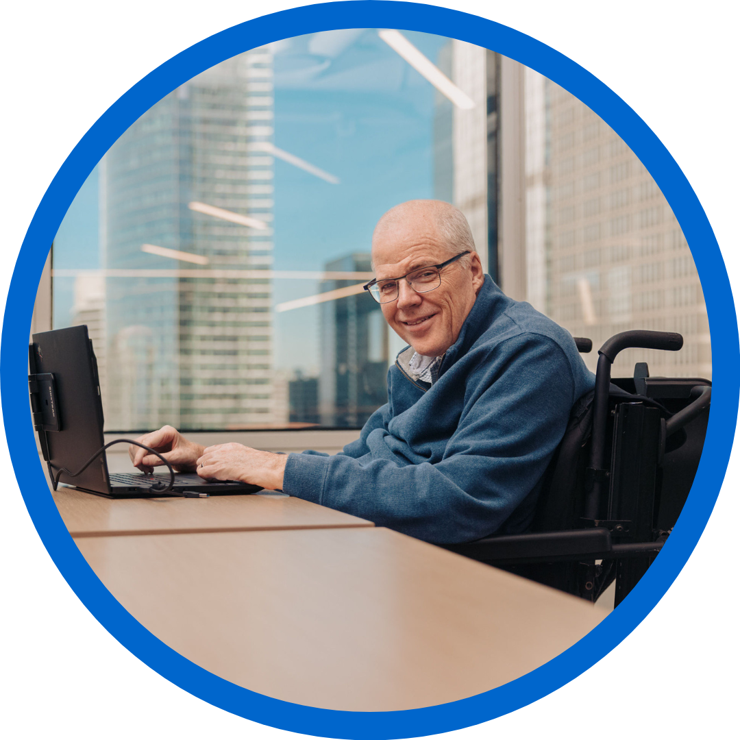 Mike smiles as he types on a keyboard in an office. He is an older man who uses a power wheelchair and is wearing glasses and a blue sweater.