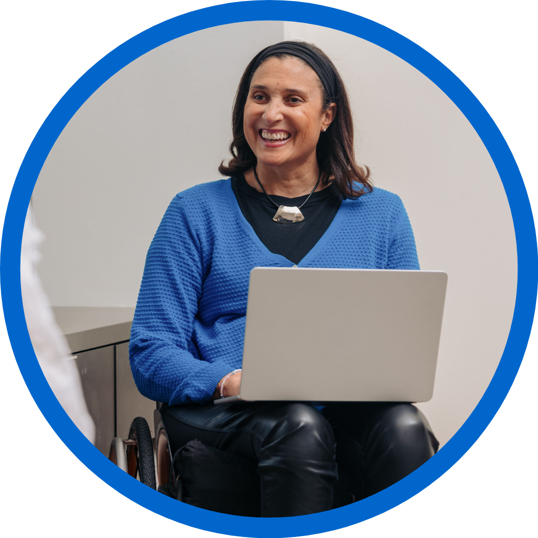Tricia smiles as she types on a laptop. She has tan skin, shoulder length brown hair, and uses a wheelchair. She wears black leather pants and a blue sweater. 