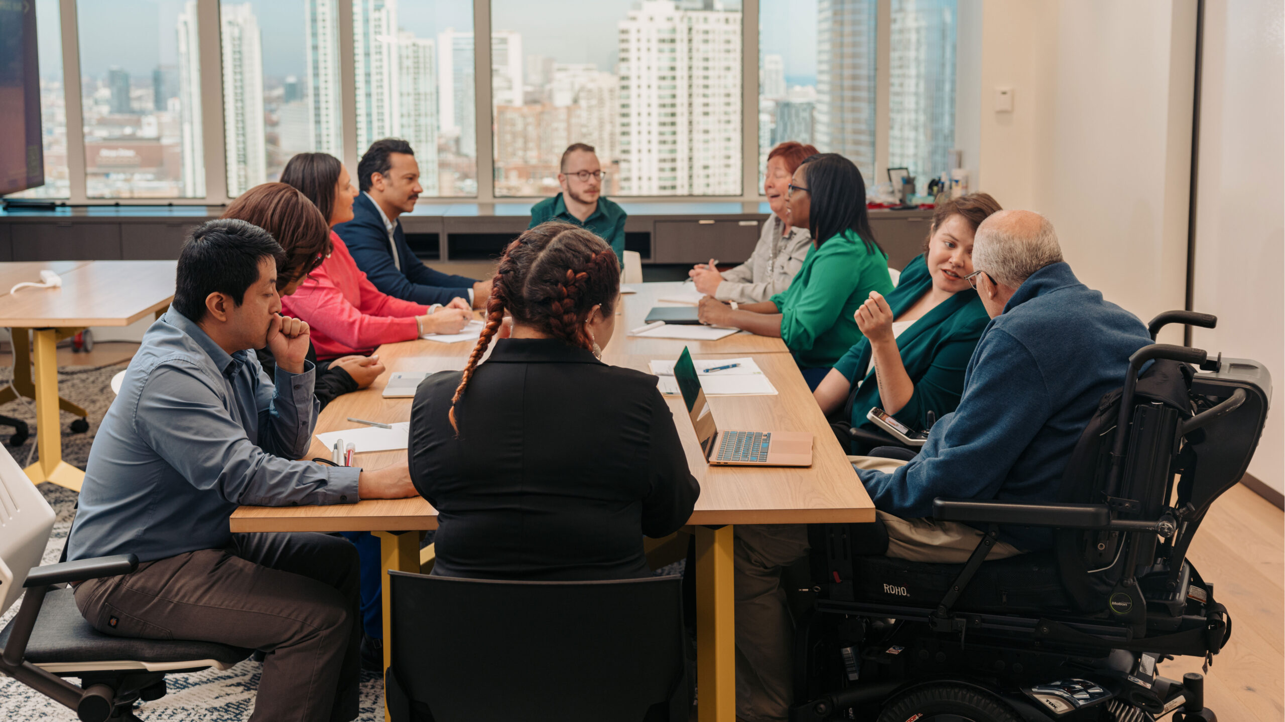 A group of diverse colleagues with disabilities gather around a conference table with the skyline showing in the background.