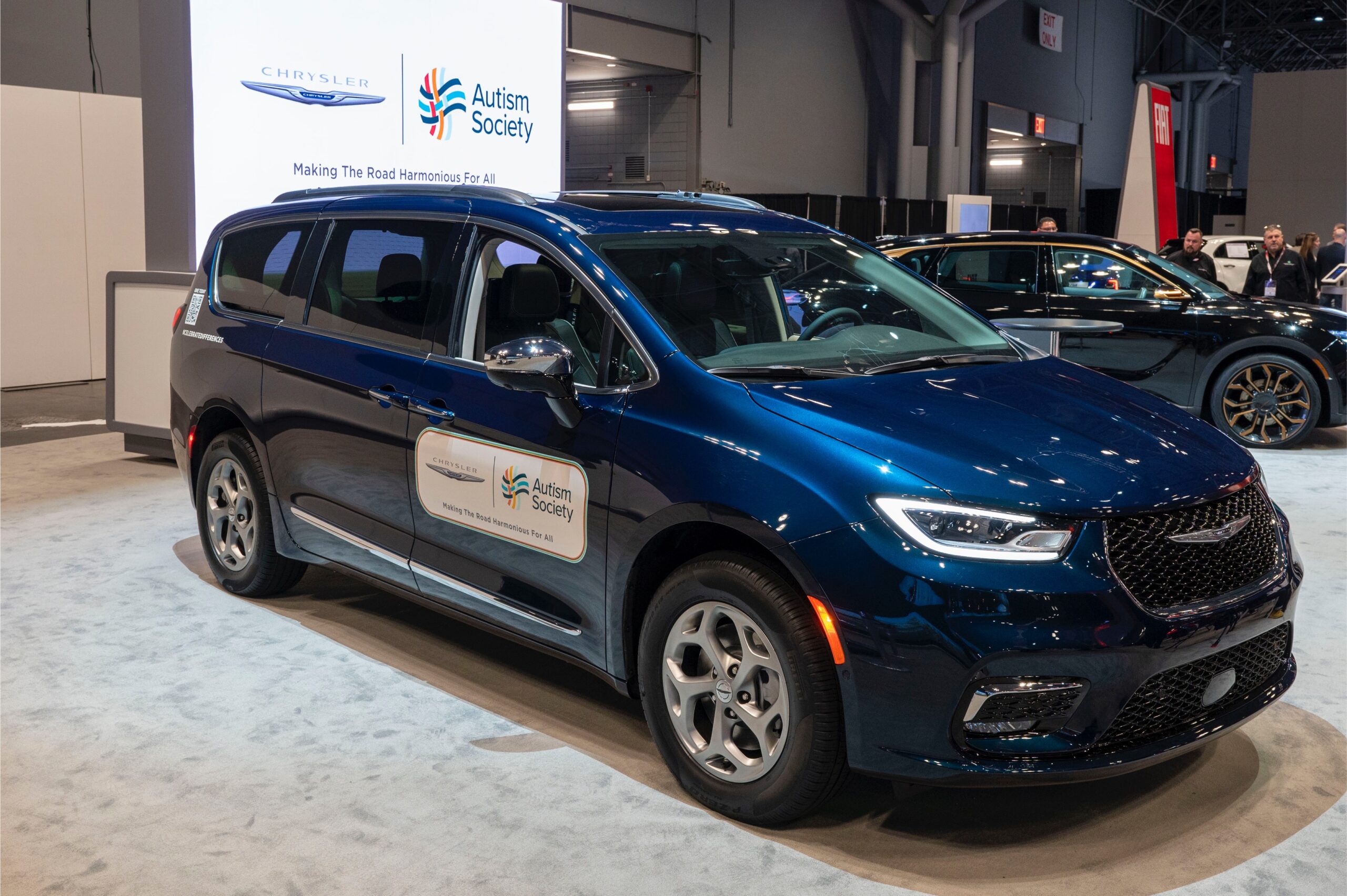 A blue Chrysler Pacifica minivan parked in a showroom. A white banner with the Autism Society and Chrysler brand logos is displayed behind the vehicle.