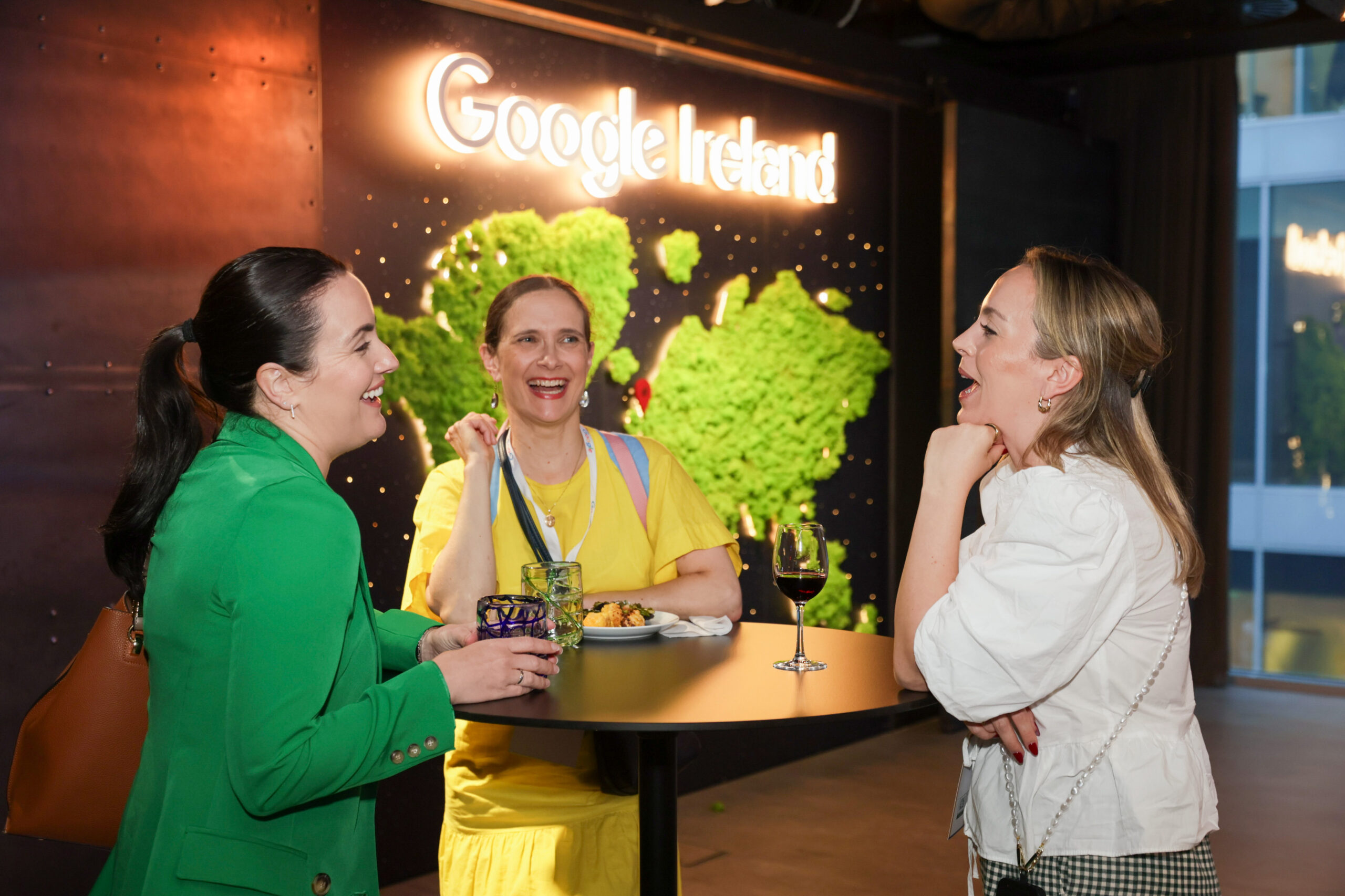 Three colleagues are laughing and conversing at a reception table in front of a Google Ireland global wall installation.