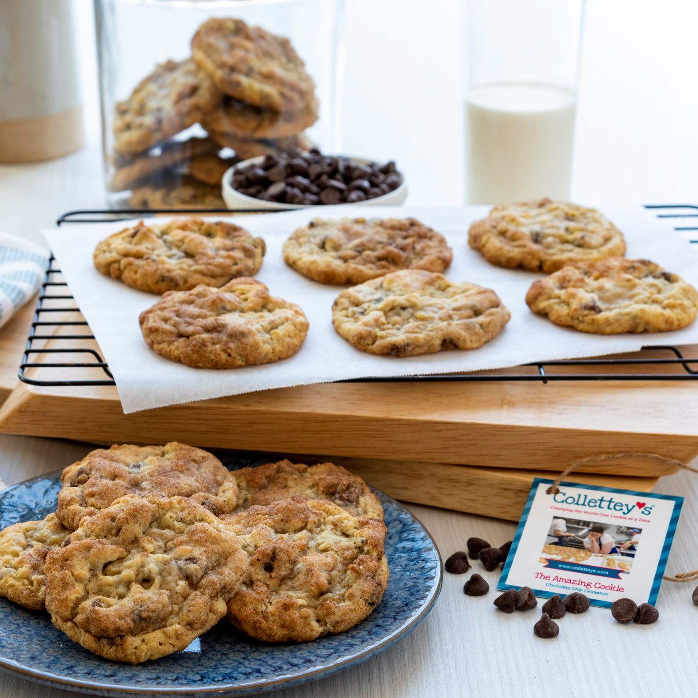 Array of chocolate chip cookies on a plate and parchment paper, with kitchen items and glass of milk nearby.