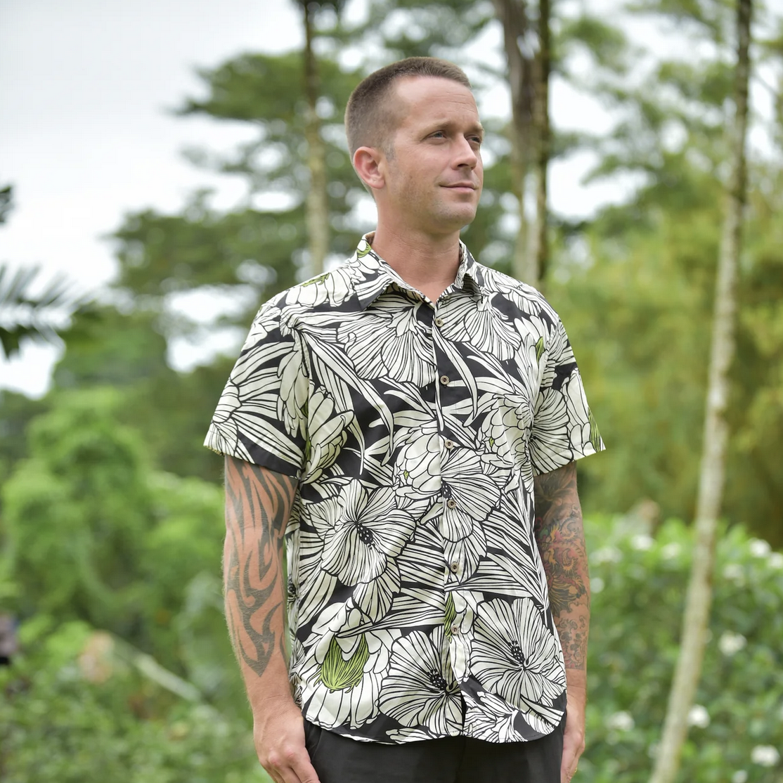 Man wears a short sleeve Coradorables Hawaiian short sleeve shirt against a tropical landscape.