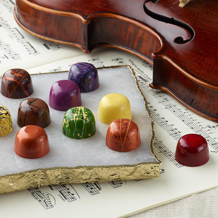 Array of colorful chocolate truffles on a stone tray next to a violin and sheet music.