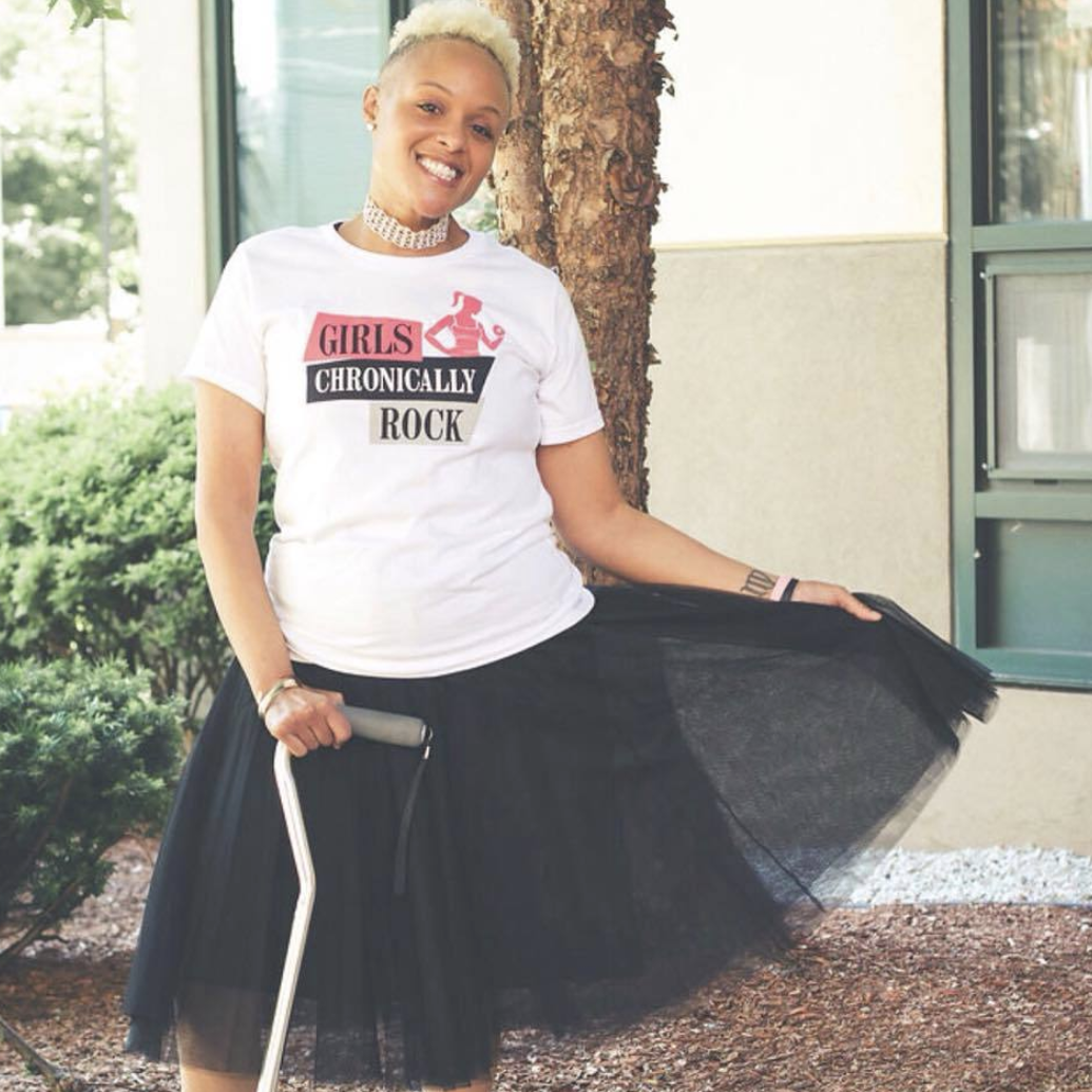 Woman wearing a Girls Chronically Rock white t-shirt, showing off her skirt and mobility aid.