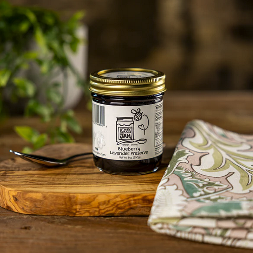 Jar of Blueberry Lavender preserve in a glass jar on top of a wooden board next to utensils and napkins.